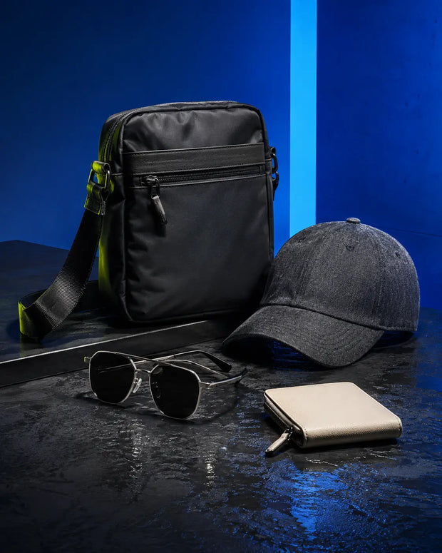 Unbranded black crossbody bag, charcoal cap, sunglasses and cream wallet in a blue studio setting